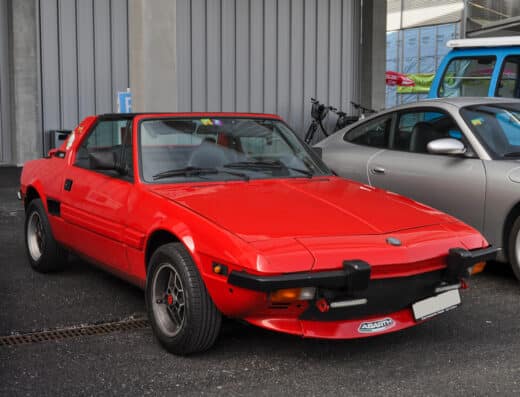 Roter klassischer Sportwagen in einer hellen Ausstellungshalle, seitlich aufgenommen, mit geöffnetem Dach und eleganten Linien.